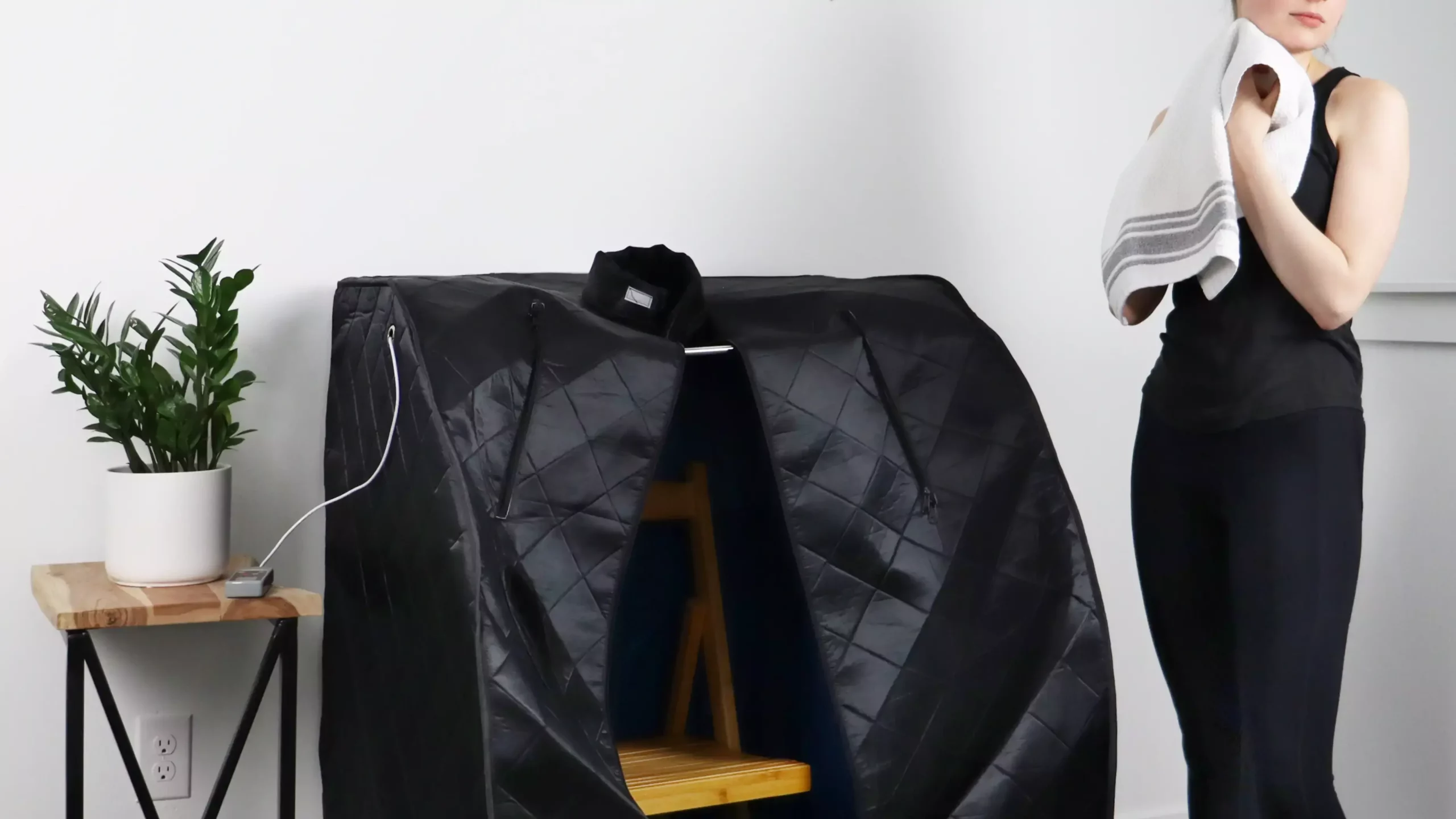Woman holding a towel standing next to an open, black ULtraLux Infrared Foldaway Sauna.